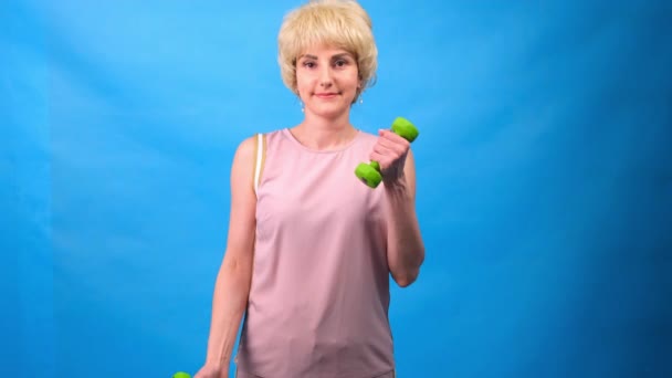 Divertente donna in una parrucca con i capelli bianchi con i manubri verdi nelle sue mani facendo esercizi su uno sfondo blu — Video Stock