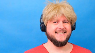 Peruklu, pembe tişörtlü garip şişman adam kulaklıkla müzik dinliyor ve mavi arka planda dans ediyor.