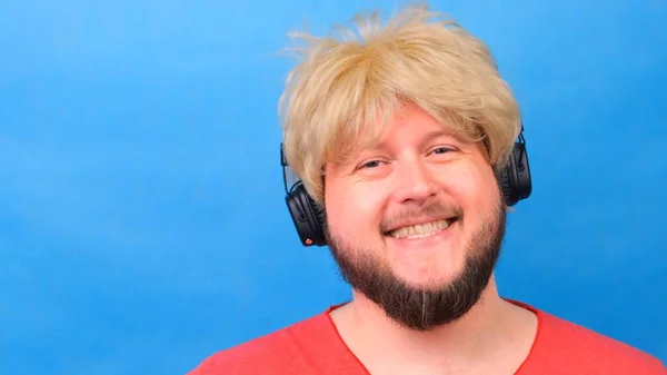 Riendo hombre gordo raro en una peluca y una camiseta rosa en sus manos escucha música en los auriculares y baila sobre un fondo azul — Foto de Stock