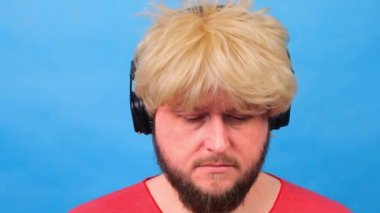 Peruklu, pembe tişörtlü, üzgün, şişman adam kulaklıkla müzik dinliyor ve mavi arka planda dans ediyor.