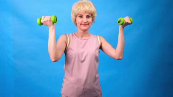 Retrato de uma mulher engraçada em uma peruca com cabelo branco, com halteres verdes em suas mãos e vestida com um traje rosa em um fundo azul — Vídeo de Stock