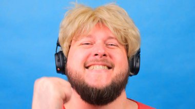 Peruklu, pembe tişörtlü garip şişman adam kulaklıkla müzik dinliyor ve mavi arka planda dans ediyor.