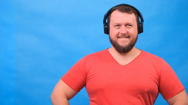 Bearded funny smiling cute fat man in a pink T-shirt listens to music and dances on a blue background, close-up, copy space. — Stock Photo, Image