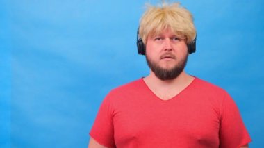 Peruklu, pembe tişörtlü, üzgün, şişman adam kulaklıkla müzik dinliyor ve mavi arka planda dans ediyor.
