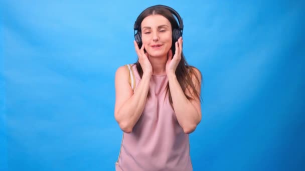 Hermosa chica de energía con auriculares escuchando música sobre un fondo azul en el estudio — Vídeos de Stock