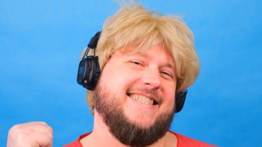 Peruklu, pembe tişörtlü garip şişman adam kulaklıkla müzik dinliyor ve mavi arka planda dans ediyor.