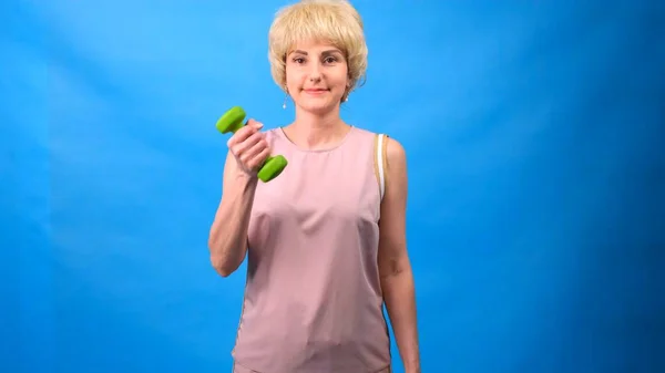 Porträt einer lustigen Frau mit Perücke, weißem Haar, grünen Hanteln in den Händen und einem rosafarbenen Trainingsanzug auf blauem Hintergrund — Stockfoto