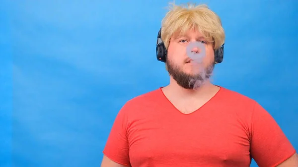 Hombre gordo raro en una peluca y una camiseta rosa en sus auriculares exhala humo de cigarrillo, fuma un vapor sobre un fondo azul . — Foto de Stock