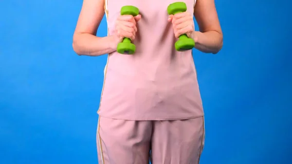 Žena v růžovém sportovním obleku s činkami v rukou dělá cvičení na modrém pozadí. Koncept sportu, stravy a hubnutí. — Stock fotografie