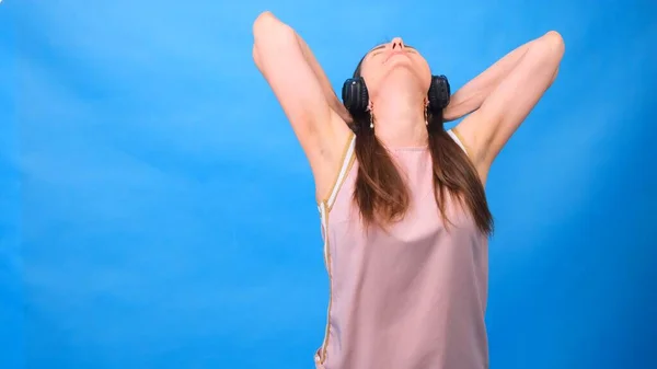 Mooie vrouw die vrolijk naar muziek luistert met een koptelefoon, poserend op een blauwe muur met vrije ruimte. — Stockfoto