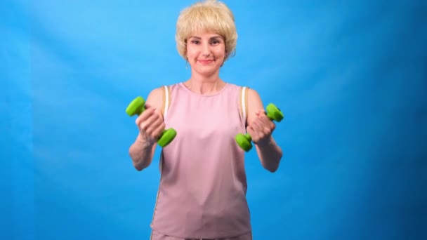 Porträt einer lustigen Frau mit Perücke, weißem Haar, grünen Hanteln in den Händen und einem rosafarbenen Trainingsanzug auf blauem Hintergrund — Stockvideo