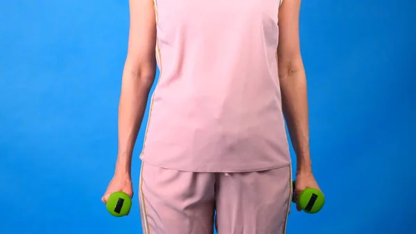 Žena v růžovém sportovním obleku s činkami v rukou dělá cvičení na modrém pozadí. Koncept sportu, stravy a hubnutí. — Stock fotografie