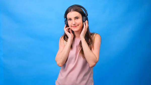 Beautiful woman joyfully listening to music using headphones, posing on a blue wall with free space. — Stock Photo, Image