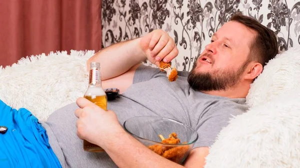 Ο χοντρός πίνει μπύρα και τρώει ανθυγιεινά φτερά κοτόπουλου, βαριέται μπροστά στην τηλεόραση στον καναπέ. Η έννοια του υποσιτισμού, καραντίνα στο σπίτι, αλκοολισμός. — Φωτογραφία Αρχείου