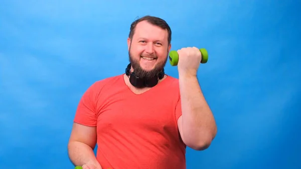 Gordo fantasia homem em um rosa t-shirt com halteres engraçado fazendo exercícios em um fundo azul . — Fotografia de Stock
