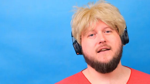 Hombre gordo raro triste en una peluca y una camiseta rosa en sus manos escucha música en los auriculares y baila sobre un fondo azul — Foto de Stock