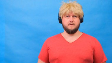 Peruklu, pembe tişörtlü, üzgün, şişman adam kulaklıkla müzik dinliyor ve mavi arka planda dans ediyor.