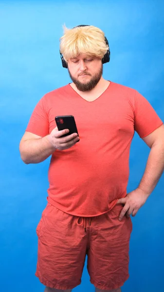 Divertido triste gordito freak hombre en auriculares y una peluca en una camiseta rosa mira en un teléfono inteligente sobre un fondo azul, vertical — Foto de Stock