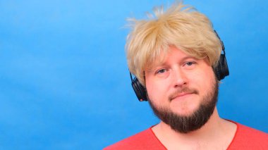 Peruklu, pembe tişörtlü, üzgün, şişman adam kulaklıkla müzik dinliyor ve mavi arka planda dans ediyor.