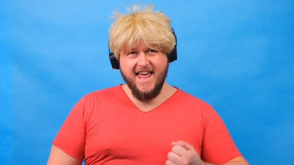 Laughing freaky fat man in a wig and a pink T-shirt in his hands listens to music on headphones and dances on a blue background — Stock Photo, Image