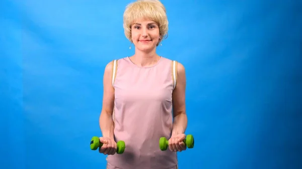 Lustige Frau in Perücke mit weißem Haar und grünen Hanteln in den Händen, die Übungen auf blauem Hintergrund machen — Stockfoto