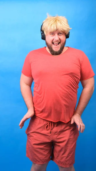 Engraçado homem aberração gordinha em fones de ouvido e uma peruca em uma camiseta rosa dança e ri em um fundo azul, vertical — Fotografia de Stock