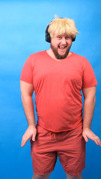 Engraçado homem aberração gordinha em fones de ouvido e uma peruca em uma camiseta rosa dança e ri em um fundo azul, vertical — Fotografia de Stock