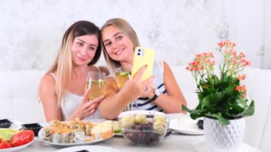  Arkadaşlık ve parti konsepti. İki güzel kız arkadaş şarap içip akıllı telefondan özçekim yapıyorlar. Sofrada rahat bir mutfakta, seçici bir şekilde.