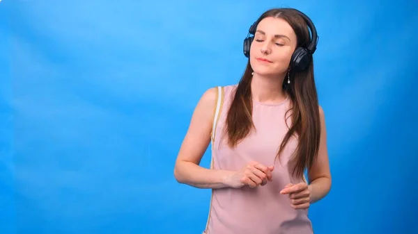 Mooie vrouw die vrolijk naar muziek luistert met een koptelefoon, poserend op een blauwe muur met vrije ruimte. — Stockfoto