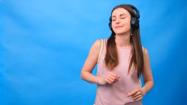 Hermosa mujer escuchando alegremente música usando auriculares, posando en una pared azul con espacio libre. — Foto de Stock