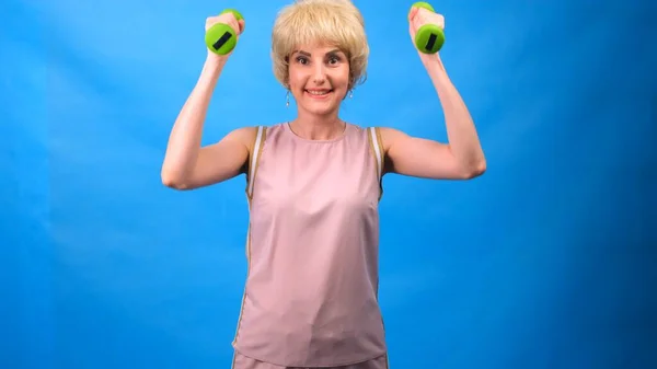 Porträt einer lustigen Frau mit Perücke, weißem Haar, grünen Hanteln in den Händen und einem rosafarbenen Trainingsanzug auf blauem Hintergrund — Stockfoto