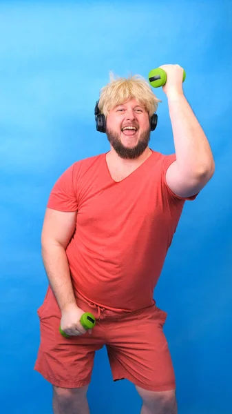 Engraçado homem aberração gordinha em fones de ouvido e uma peruca em uma camiseta rosa está envolvida em halteres verdes e ri em um fundo azul, vertical — Fotografia de Stock
