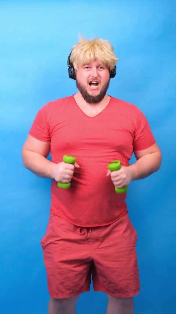 Grappige mollige freak man in hoofdtelefoon en een pruik in een roze t-shirt is bezig met groene halters en lacht op een blauwe achtergrond, verticale — Stockvideo