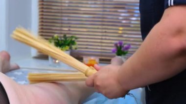 Masaj terapisti, masaj masasında elinde bambu çubuğuyla oturan şişman bir kadına selülit karşıtı bacak masajı yapıyor. Spa tedavileri. Sağlık ve güzellik, diyet