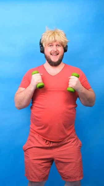 Engraçado homem aberração gordinha em fones de ouvido e uma peruca em uma camiseta rosa está envolvida em halteres verdes e ri em um fundo azul, vertical — Fotografia de Stock