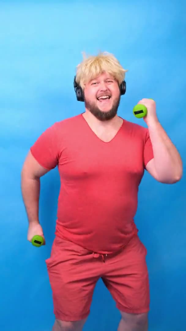 Grappige mollige freak man in hoofdtelefoon en een pruik in een roze t-shirt is bezig met groene halters en lacht op een blauwe achtergrond, verticale — Stockvideo