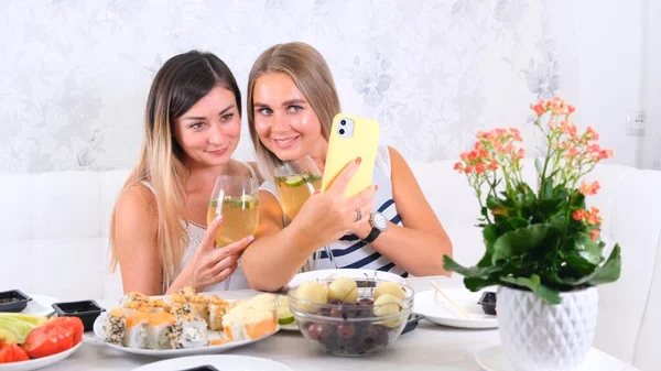 Friendship and party concept - two young beautiful girlfriends drink wine and take selfie on smartphone in a cozy kitchen at the table, selective focus — Stock Photo, Image