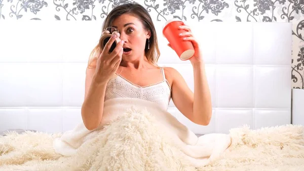 Attractive young woman gets up in bed with a cup of coffee and an alarm clock. The woman overslept and is late, reacting with horror at the time.