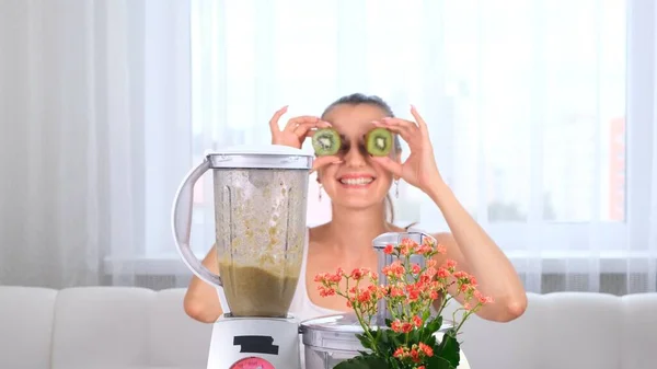 Boldog fiatal nő készül finom tápláló smoothie a turmixgépben. vicces nő csinál szemek kiwi. — Stock Fotó