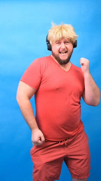 Engraçado homem aberração gordinha em fones de ouvido e uma peruca em uma camiseta rosa dança e ri em um fundo azul, vertical — Fotografia de Stock