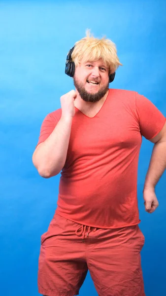 Engraçado homem aberração gordinha em fones de ouvido e uma peruca em uma camiseta rosa dança e ri em um fundo azul, vertical — Fotografia de Stock