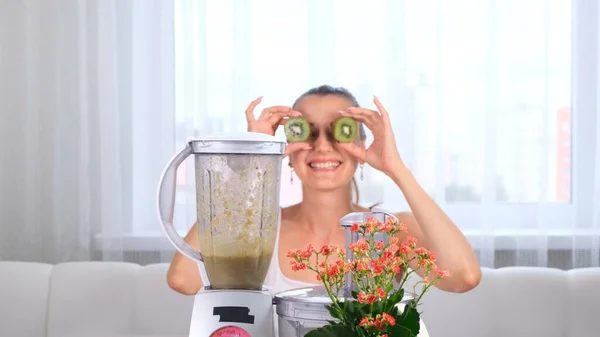 Boldog fiatal nő készül finom tápláló smoothie a turmixgépben. vicces nő csinál szemek kiwi. — Stock Fotó