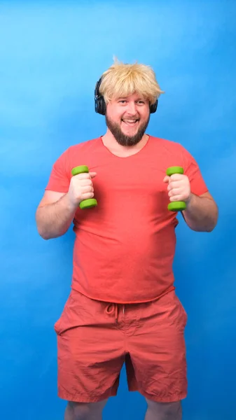 Funny chubby freak man in headphones and a wig in a pink t-shirt is engaged in green dumbbells and laughs on a blue background, vertical — Stock Photo, Image