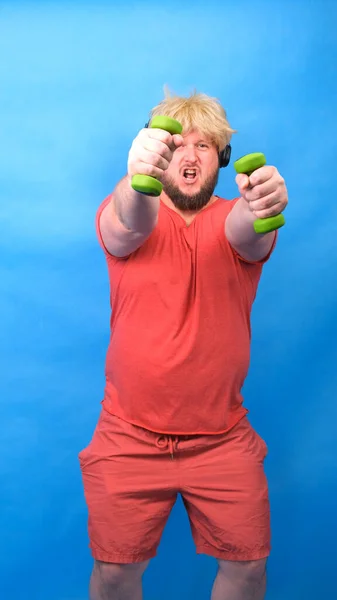 Grappige mollige freak man in hoofdtelefoon en een pruik in een roze t-shirt is bezig met groene halters en lacht op een blauwe achtergrond, verticale — Stockfoto