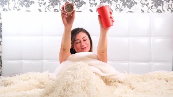 Woman rises in the bed with an alarm clock and takeaway coffee in her hands, enjoys the morning and drinks coffee. The concept of starting a new day, relaxing at the weekend, no need to rush anywhere — Stock Video