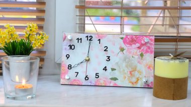 Çalar saat sabah 8 ya da akşam gösterilir. Pencerenin üzerinde renkli bir çalar saat duruyor. Mumlar ve sarı çiçeklerin yanında.