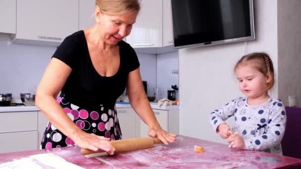 A avó ajuda a filha a enrolar a massa na cozinha para fazer biscoitos. Vovó e filha assar pizza juntos na cozinha. A menina ajuda sua mãe a distribuir a massa com um rolo. — Vídeo de Stock