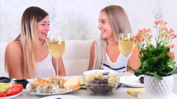 Δύο ελκυστικές κοπέλες, χαρούμενες κολλητές που διασκεδάζουν και πίνουν κρασί στο σπίτι. Δύο ποτήρια λευκό κρασί στα χέρια. Κορίτσια που διασκεδάζουν μαζί κουτσομπολεύοντας. — Αρχείο Βίντεο