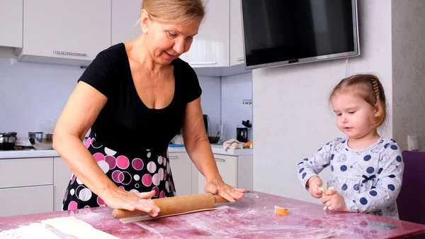 Grandma helps her daughter roll the dough in the kitchen to bake cookies. Grandma and daughter bake pizza together in the kitchen. The girl helps her mother roll out the dough with a rolling pin.