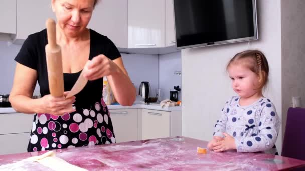 Oma helpt haar dochter het deeg in de keuken te rollen om koekjes te bakken. Oma en dochter bakken samen pizza in de keuken. Het meisje helpt haar moeder het deeg uit te rollen met een deegroller. — Stockvideo